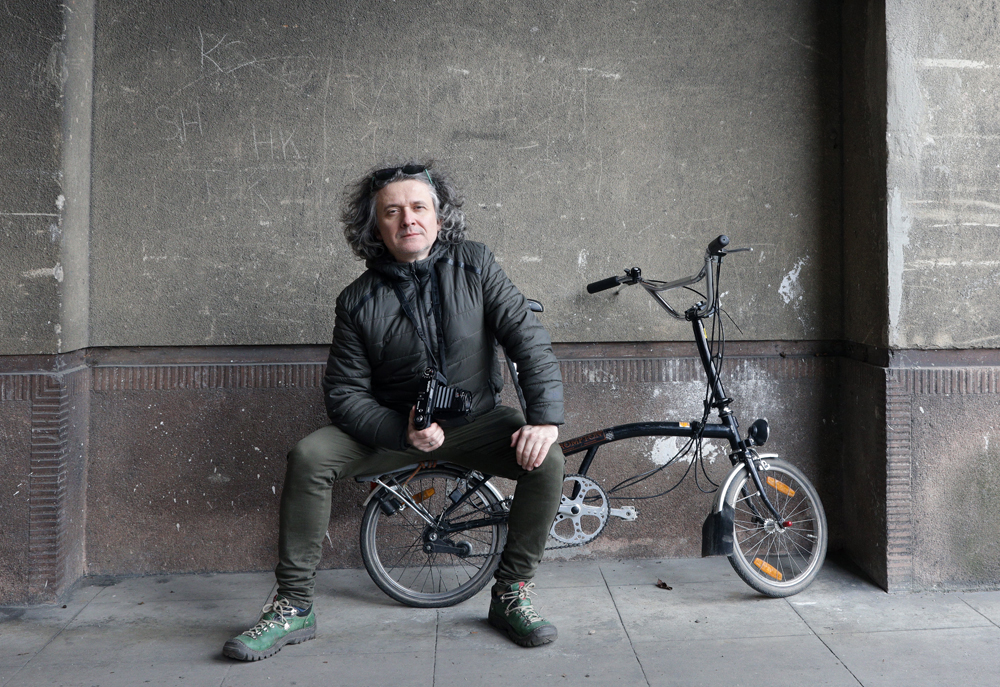 Maciej Rawluk, a middle-aged man with shoulder-length gray hair, sitting on a foldable bike with a camera in his hand