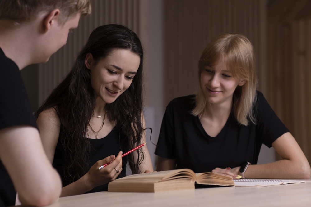 2 studentki i student siedzą przy stole, patrzą w ksiażkę i rozmawiają