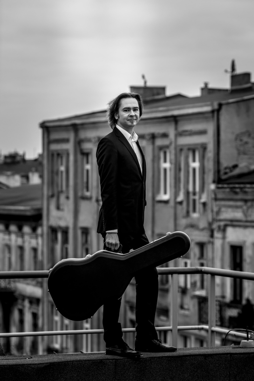 Maciej Staszewski wearing a suit and standing with a guitar on the balcony, with tenement houses visible in the background